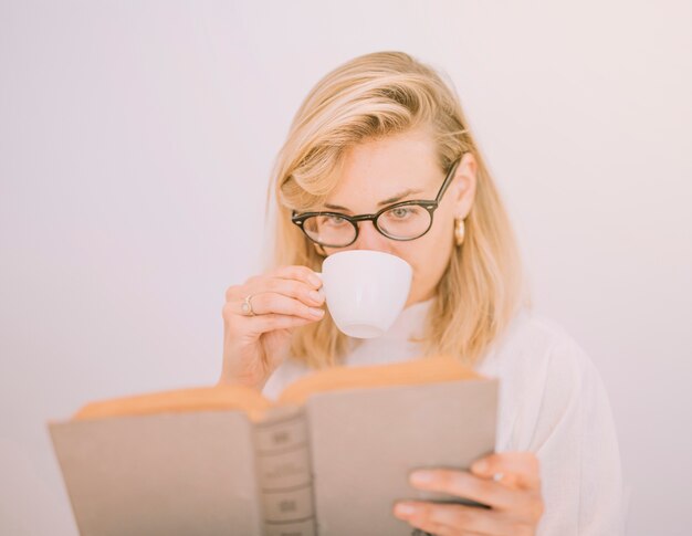 Mujer joven rubia que bebe el café mientras que lee el libro contra el fondo blanco