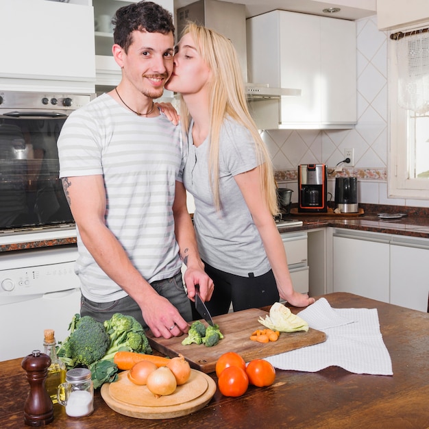 Mujer joven rubia que besa a su marido que corta la verdura en la cocina