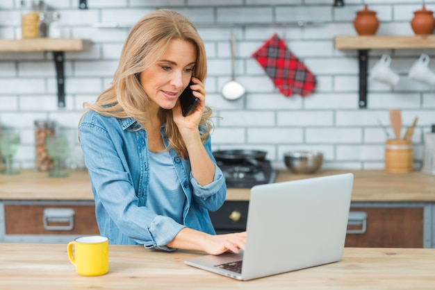 Foto gratuita mujer joven rubia que habla en el teléfono móvil usando el ordenador portátil en la tabla