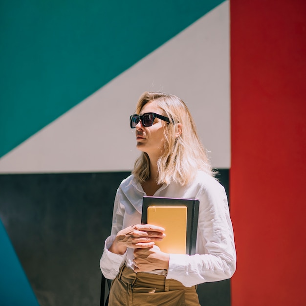 Mujer joven rubia que lleva anteojos negros que sostienen el libro en la mano que se coloca delante de e = pared colorida