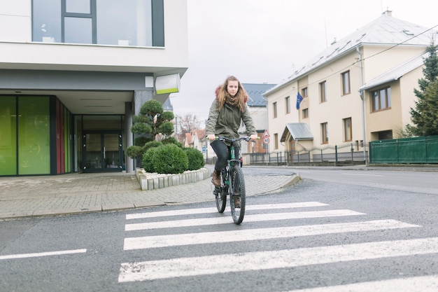 Mujer joven rubia que lleva la bicicleta superior con capucha del montar a caballo en la ciudad