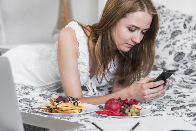 Mujer joven rubia que miente en cama con las placas de la galleta y de las frutas usando smartphone