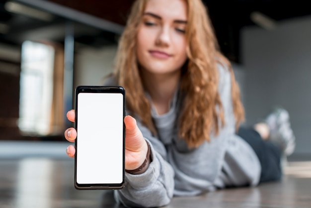 Mujer joven rubia que miente en el piso que muestra el teléfono móvil con la pantalla de visualización blanca