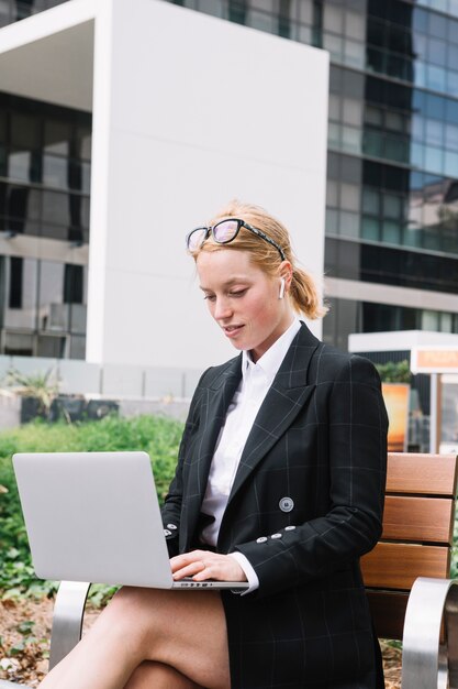 Mujer joven rubia que se sienta en banco usando la computadora portátil