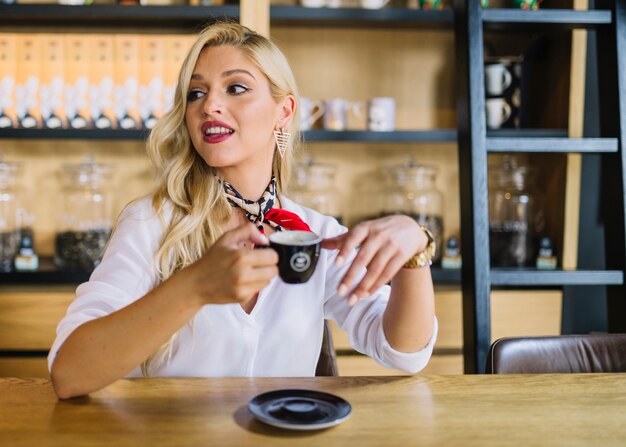 Mujer joven rubia que se sienta en el café que sostiene la taza de café que mira lejos