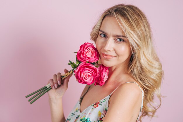Mujer joven rubia que sostiene rosas disponibles contra fondo rosado