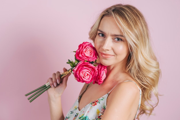 Mujer joven rubia que sostiene rosas disponibles contra fondo rosado