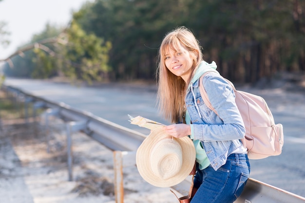Mujer joven rubia que sostiene el sombrero y el mapa que se colocan cerca del camino con su mochila que mira la cámara