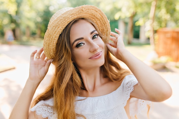 Mujer joven rubia refinada sonriendo suavemente y sosteniendo el sombrero de paja vintage. Retrato de primer plano de linda chica de buen humor posando con placer en el parque.