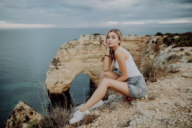 Mujer joven rubia sentada en la cima de las rocas junto al océano.