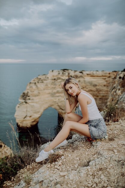 Mujer joven rubia sentada en la cima de las rocas junto al océano.