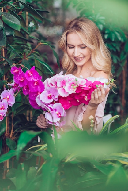 Mujer joven rubia sonriente que se coloca delante de las plantas verdes que miran las flores rosadas exóticas de la orquídea