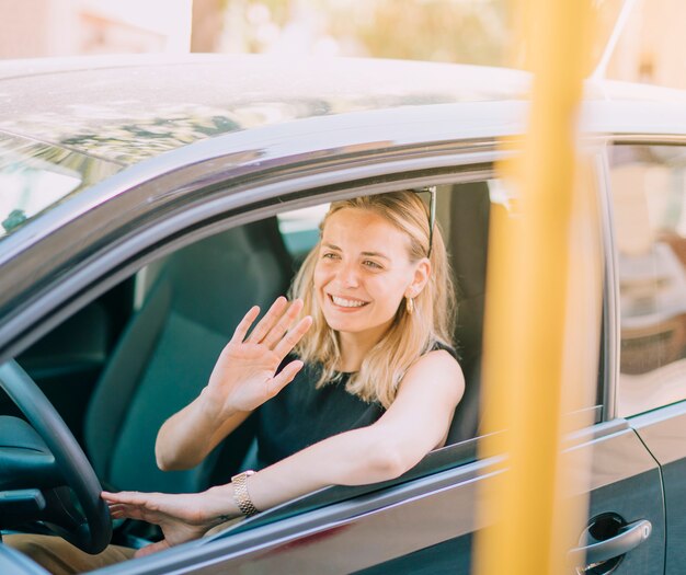 Mujer joven rubia sonriente que conduce el coche que agita su mano