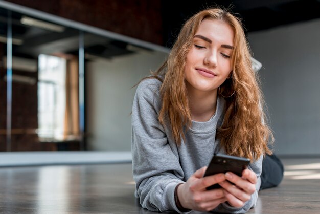 Mujer joven rubia sonriente que miente en piso usando el teléfono móvil
