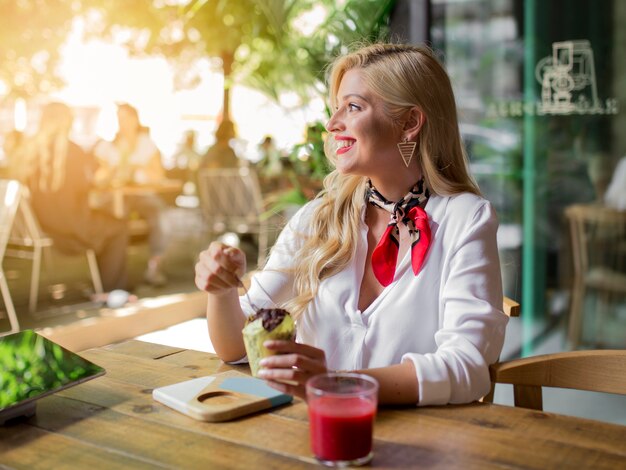 Mujer joven rubia sonriente que se sienta en el café que come el mollete