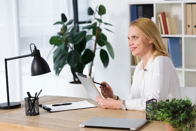 Mujer joven rubia sonriente que se sienta en el lugar de trabajo usando la tableta digital