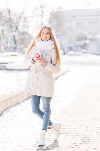 Mujer joven rubia sonriente que sostiene la taza de café disponible que se coloca en la calle en invierno