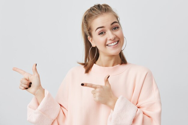 Mujer joven rubia sonriente en sudadera rosa que señala en el espacio de la copia para su anuncio o texto. Chica positiva con una sonrisa demoníaca sus dientes blancos anunciando algo