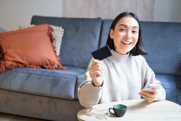 Mujer joven en la sala de estar mostrando su tarjeta de crédito y usando un teléfono inteligente para pagar el pedido en línea o comprar