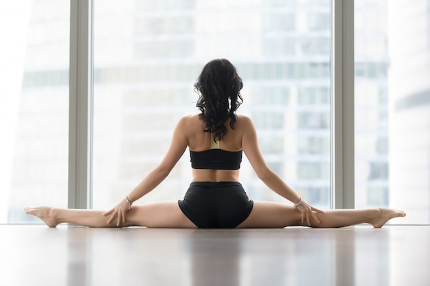 Mujer joven en Samakonasana posa contra la ventana del piso, vista trasera