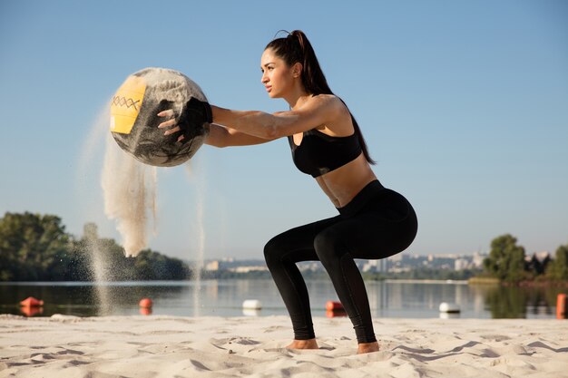 Mujer joven sana haciendo sentadillas con la pelota en la playa.