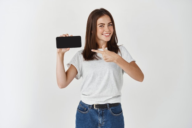 Mujer joven satisfecha que muestra la pantalla en blanco del teléfono inteligente, apuntando a la pantalla del móvil y sonriendo, recomendando la aplicación o el sitio de compras, pared blanca.