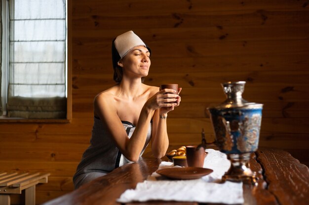 Mujer joven en una sauna con una gorra en la cabeza se sienta en una mesa y bebe té de hierbas, disfrutando de un día de bienestar