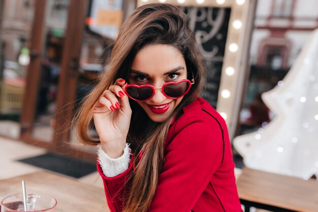 Mujer joven segura con hermoso cabello castaño mirando a la cámara a través de gafas divertidas