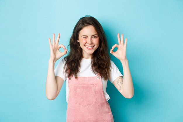 Mujer joven segura y positiva guiñando un ojo y sonriendo, mostrando señales de aprobación, decir sí, dar aprobación, elogiar el buen trabajo, de pie contra el fondo azul.