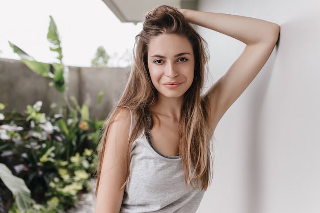 Mujer joven sensual con la expresión de la cara tranquila posando. Foto de hermosa niña con cabello largo brillante de pie frente a una gran planta verde.