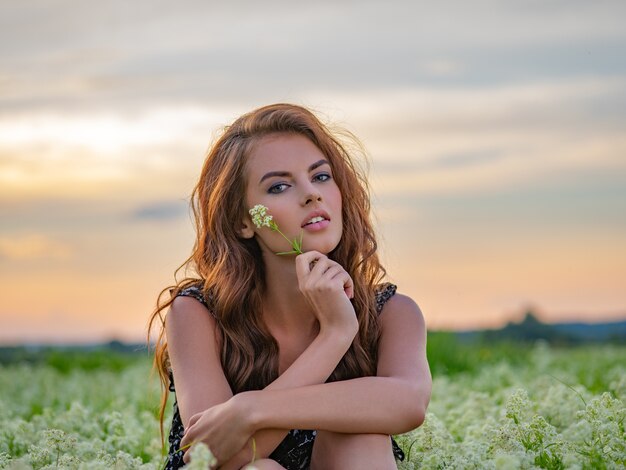 Mujer joven sentada al aire libre en un campo de flores blancas. Modelo posando en flores blancas de lavanda sosteniendo una en la mano.