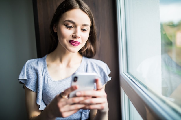 Mujer joven sentada en un alféizar de la ventana en casa y enviando mensajes de texto en su teléfono comunicación femenina mirando mensaje