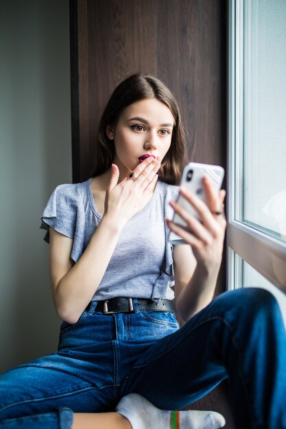 Mujer joven sentada en el alféizar de una ventana en casa y enviando mensajes de texto en su teléfono comunicación mensaje femenino