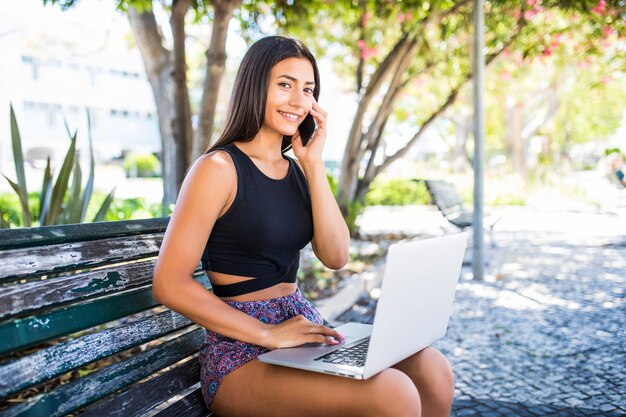 Mujer joven sentada en un banco, hablando por teléfono inteligente, trabajando en la computadora portátil al aire libre.