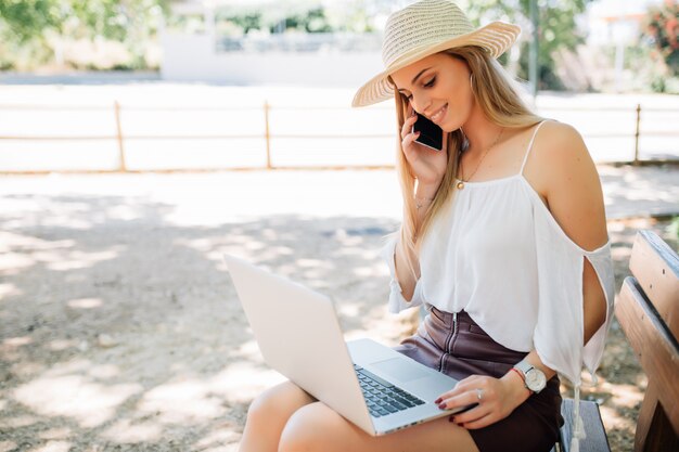 Mujer joven sentada en un banco en el parque hablando por teléfono celular y usando la computadora portátil.