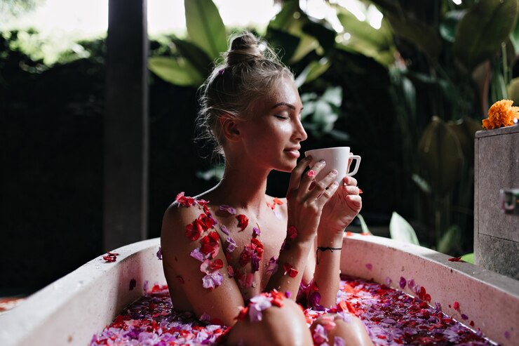 mujer joven sentada en el baño con los ojos cerrados y bebiendo té caliente. Retrato de niña magnífica con cabello rubio haciendo spa y disfrutando de un café.