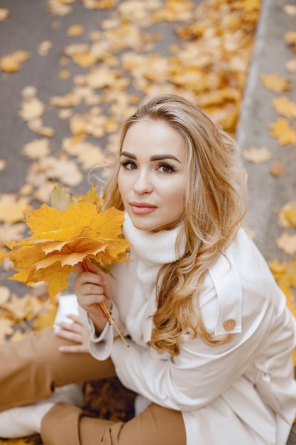 Mujer joven sentada en un bordillo en el bosque de otoño. Mujer rubia sosteniendo hojas amarillas. Chica con abrigo beige y pantalón marrón.