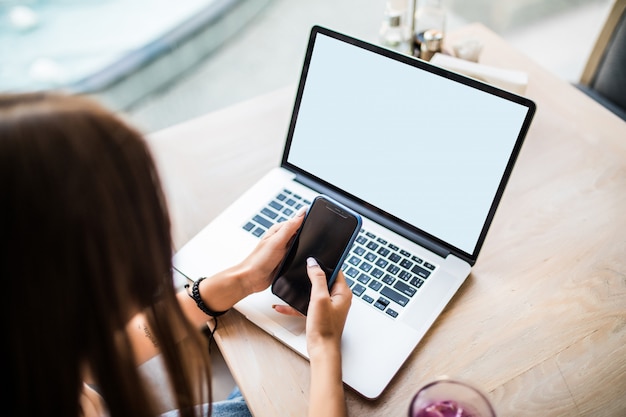 Mujer joven sentada en la cafetería en la mesa de madera con laptop, tomando café y usando el teléfono inteligente.