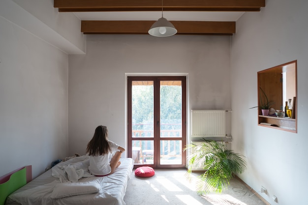 Mujer joven sentada en la cama y mirando por la ventana.