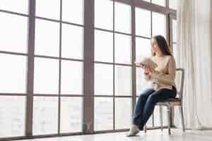 Foto gratuita mujer joven sentada cerca de la ventana leyendo el libro