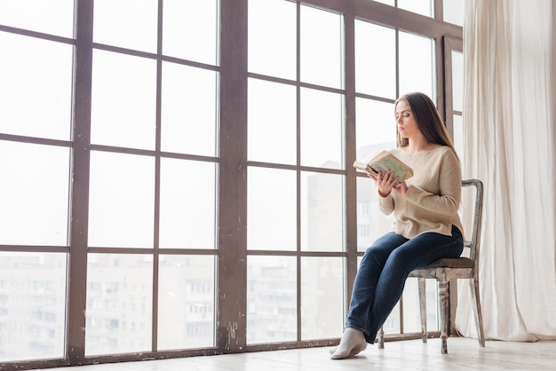 Mujer joven sentada cerca de la ventana leyendo el libro