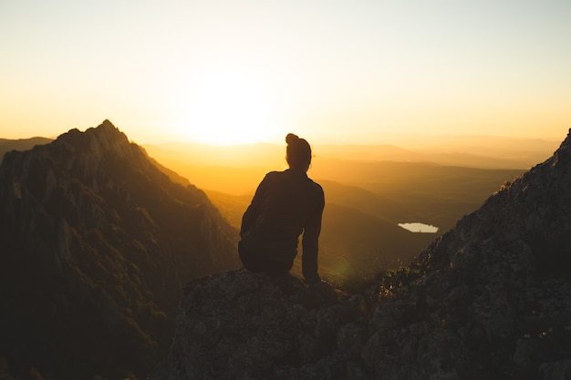 Mujer joven sentada en la cima de la montaña y disfrutando de la vista durante la puesta de sol