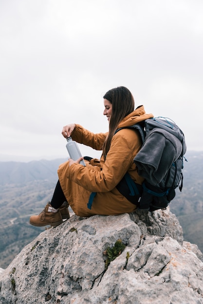 Foto gratuita mujer joven sentada en la cima de la montaña con su mochila sosteniendo la botella de agua