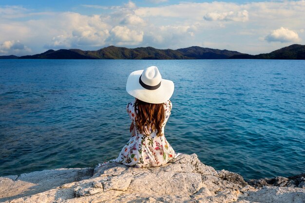 Mujer joven sentada en la cima de la roca y mirando a la orilla del mar.