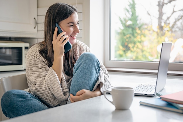 Foto gratuita una mujer joven está sentada en la cocina con un teléfono inteligente y una computadora portátil