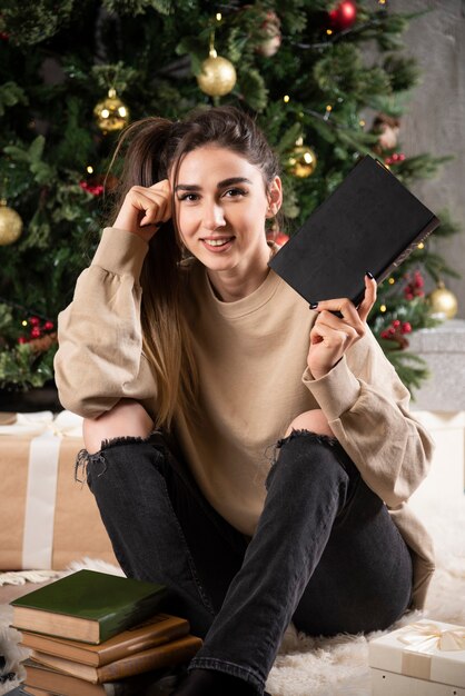 Mujer joven sentada con cuaderno negro cerca del árbol de Navidad.