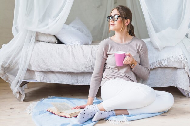 Mujer joven sentada en el dormitorio sosteniendo la taza de café con el libro
