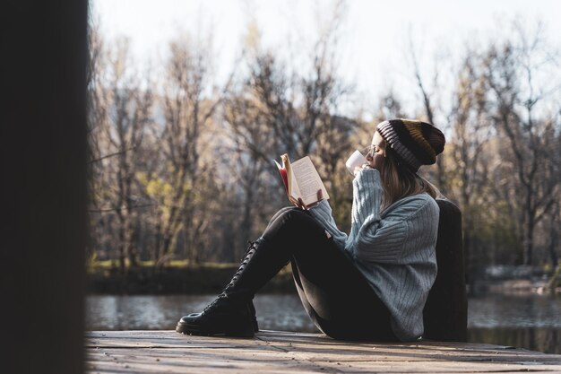 Mujer joven sentada en el embarcadero sobre el lago y leyendo un libro