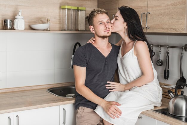 Mujer joven sentada en el mostrador de la cocina amando a su marido en sus mejillas