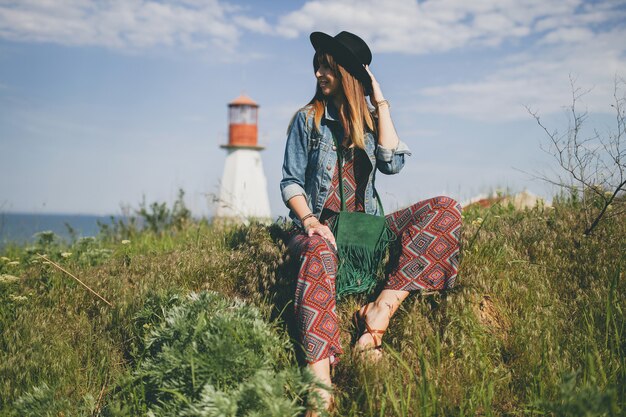 Mujer joven sentada en la naturaleza, faro, traje bohemio, chaqueta vaquera, sombrero negro, sonriente, feliz, verano, accesorios elegantes